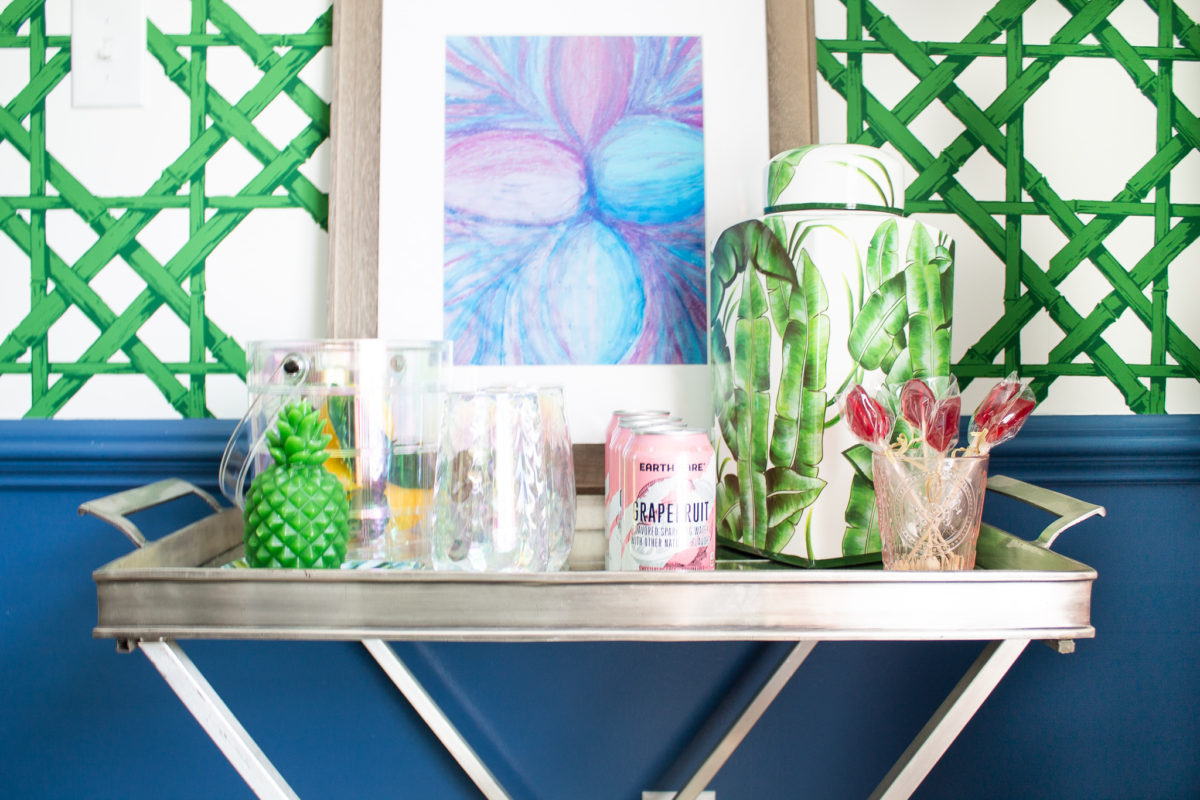 Silver bar cart in dining room against the wall with green wallpaper and artwork. The bar cart has drinks and glassware.